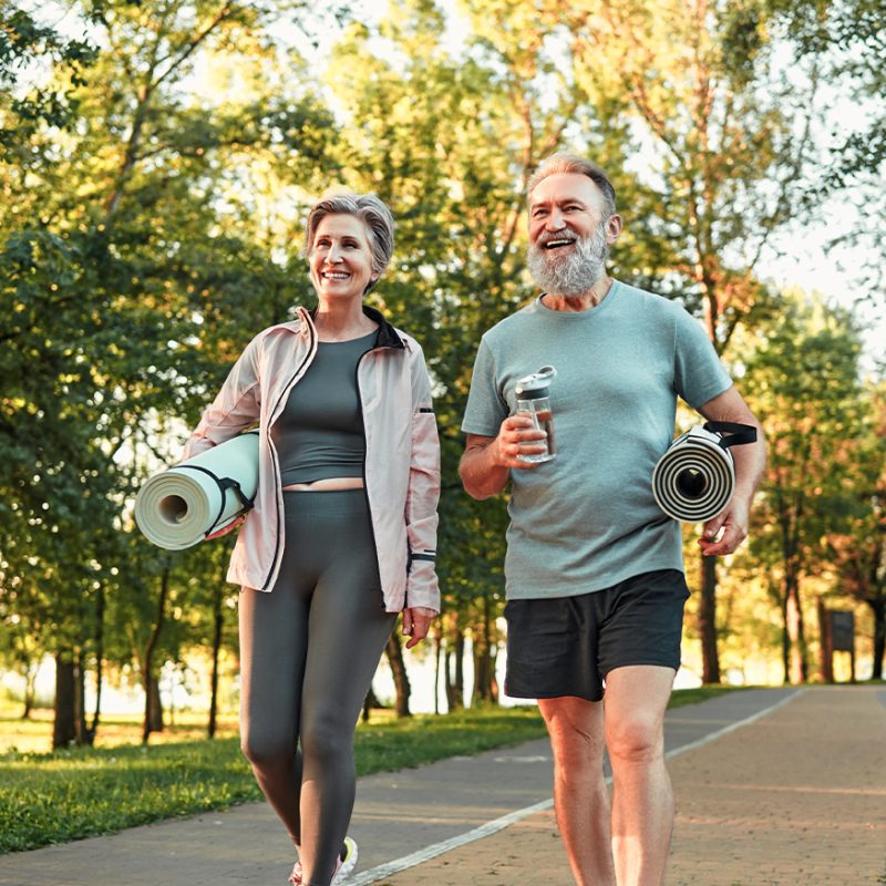 Cheerful elderly Couple walking outside in fitness gear, both holding a yoga mat.